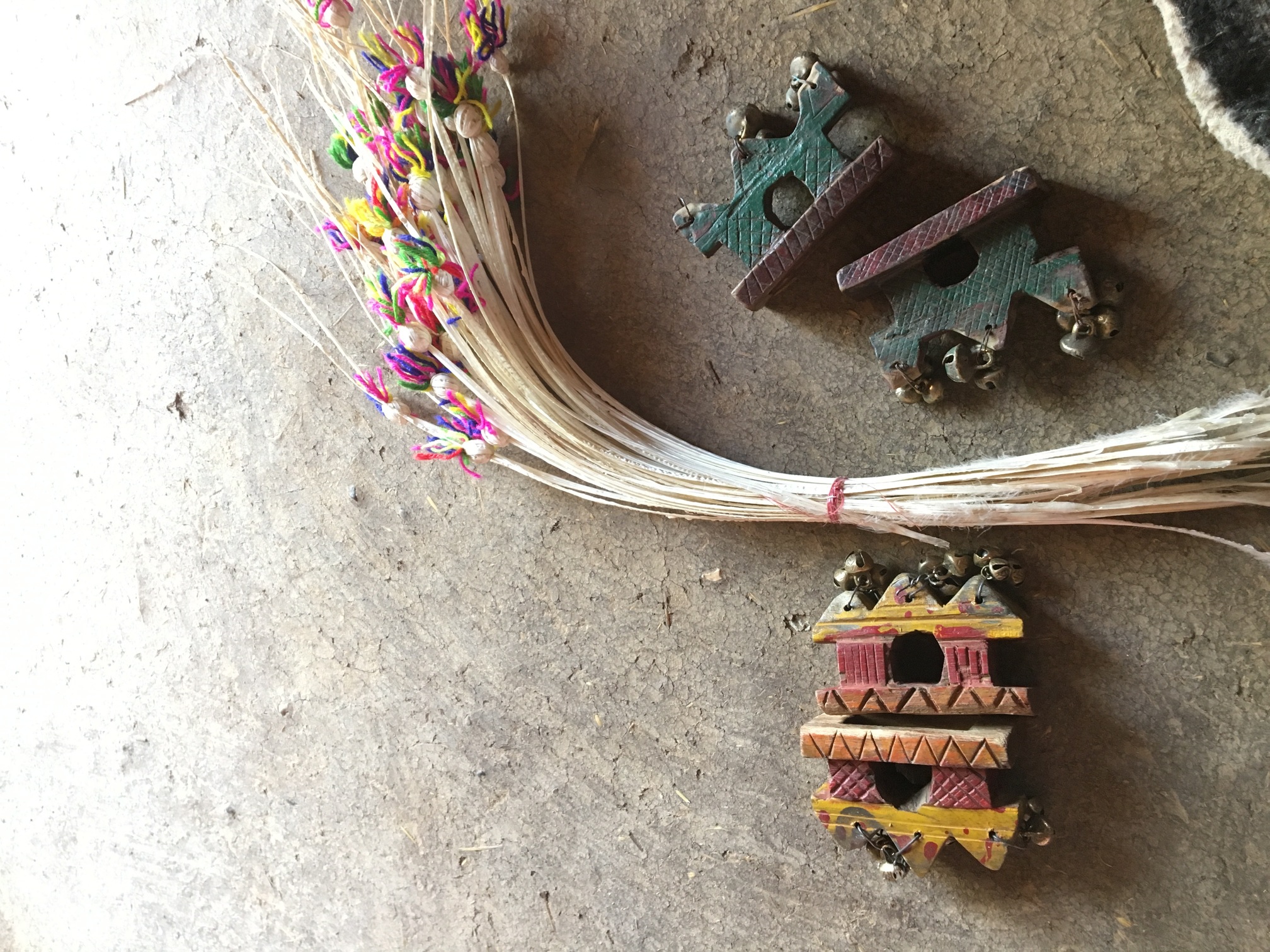 Carved wooden ornaments hung with small bells beside a bundle of reeds tied with coloured thread, on a mud floor